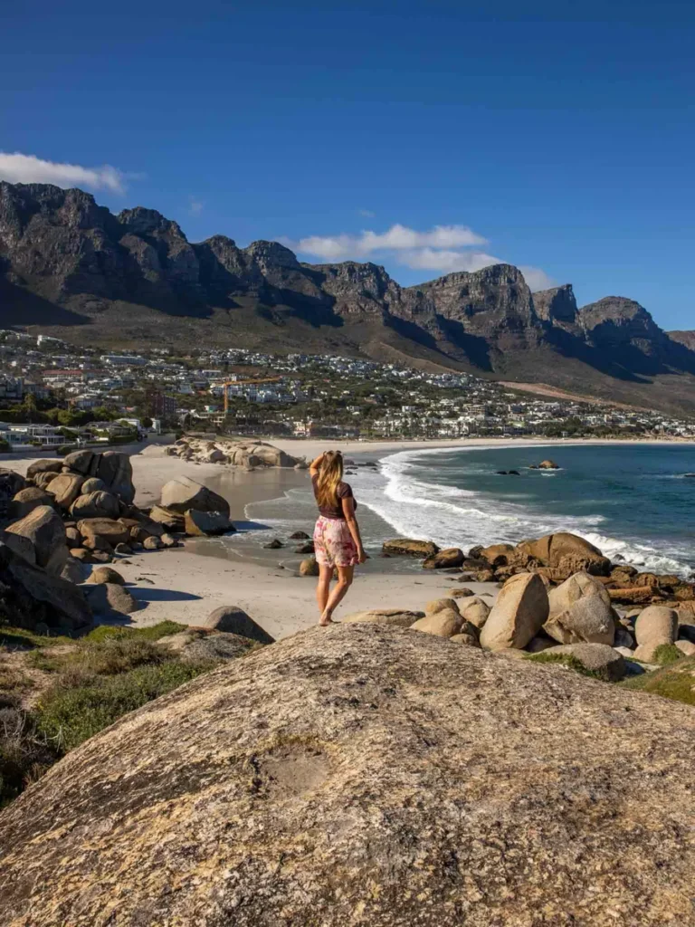 woman standing on a large rock overlooking a beautiful sandy beach with large rocks and a mountain backdrop