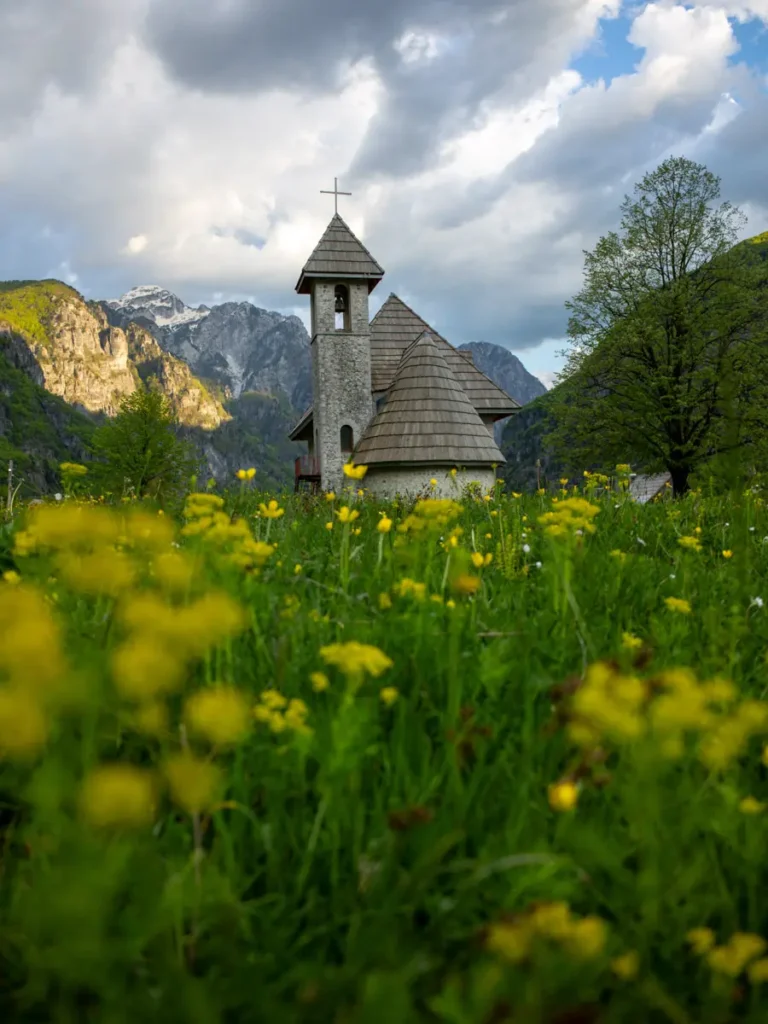 Small church with wooden roof with yellow flowers and grass in the foreground and mountains in the background in Theth, Albania