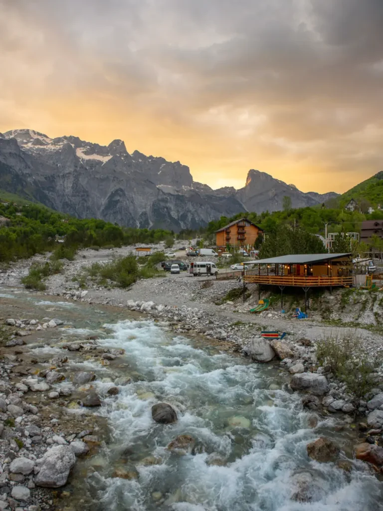 a river with rocks and mountains in the backdrop, a wooden house and a sky full of colourful clouds during sunset in Theth, Albania