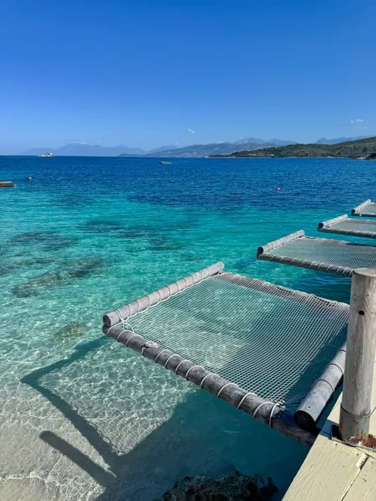 clear ocean in different shades of blue and green with a stretch of sandy beach and a few hammocks above the water