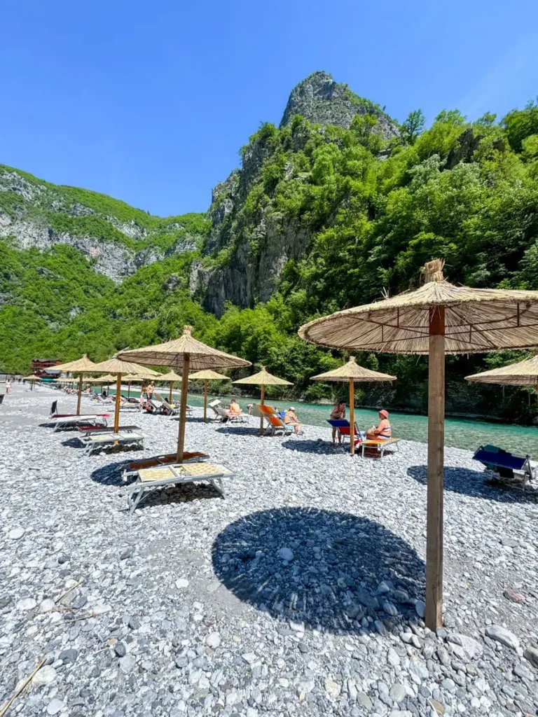 Wooden umbrellas on a riverbank at Shala River with pebbles and greenery in the distance - one of the most beautiful places in Albania