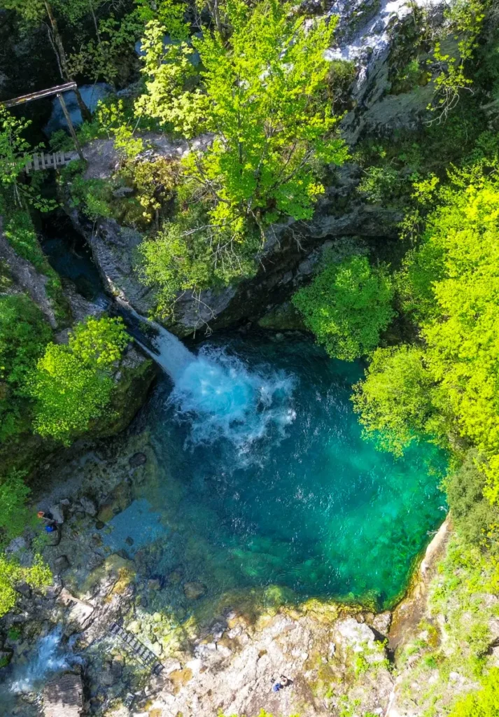 Bird's eye view of a blue spring with a little waterfall and a lot of green vegetation in Theth - a must stop on your Albania itinerary