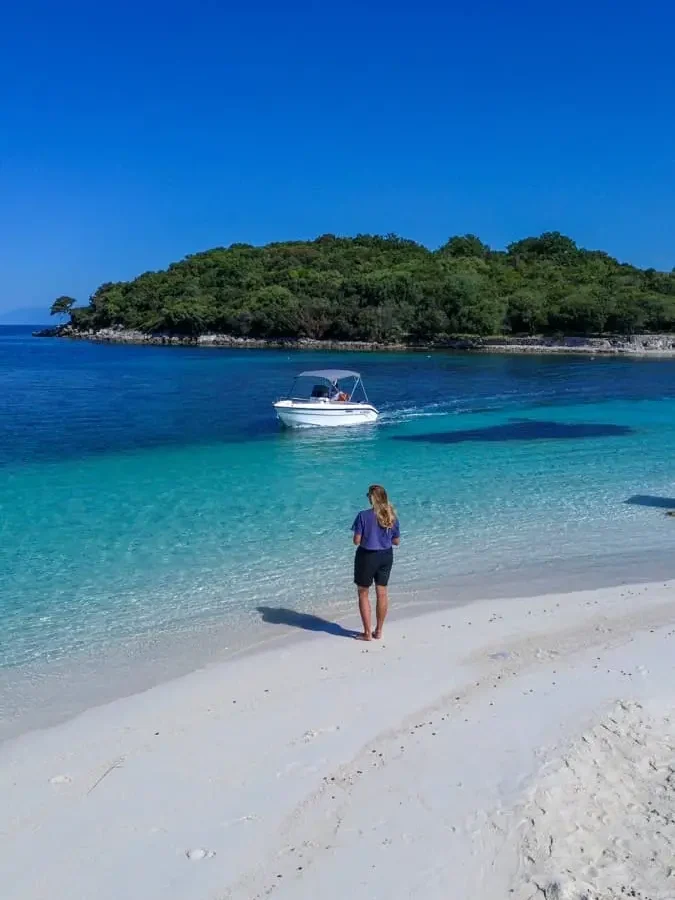 woman on a white sandy beach with crystal clear turquoise water, a small boat passing by and an island in the distance in Ksamil, Albania