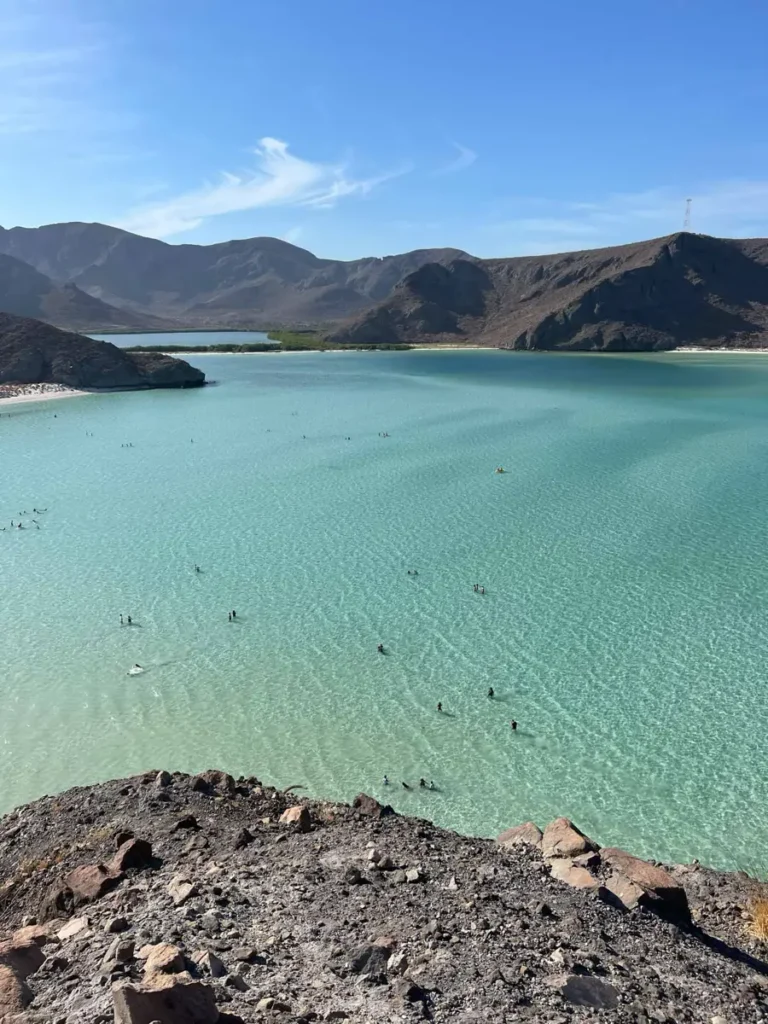 picture from above overlooking a white beach with crystal clear turquoise water with in a dry rocky hilly area with a blue sky at Playa Balandra in Baja California Sur Mexico