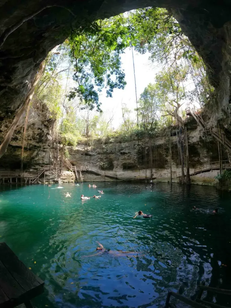 cenote in tulum mexico crystal clear blue and turquise water with rocks covered by plants with sunlight hitting the water with people swimming in the cenote