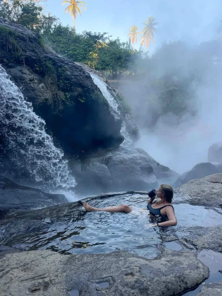 woman laying in a natural pool of a waterfall sipping on a cup of coffee with steam coming up in the back from the hot spring waterfall