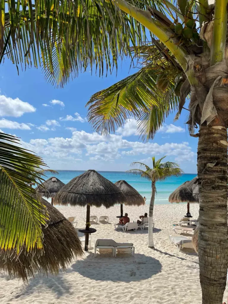white sand beach with palm trees and umbrellas made out of old palm tree leaves and lay chairs in front of the blue sea with blue sky and white clouds at Cancun