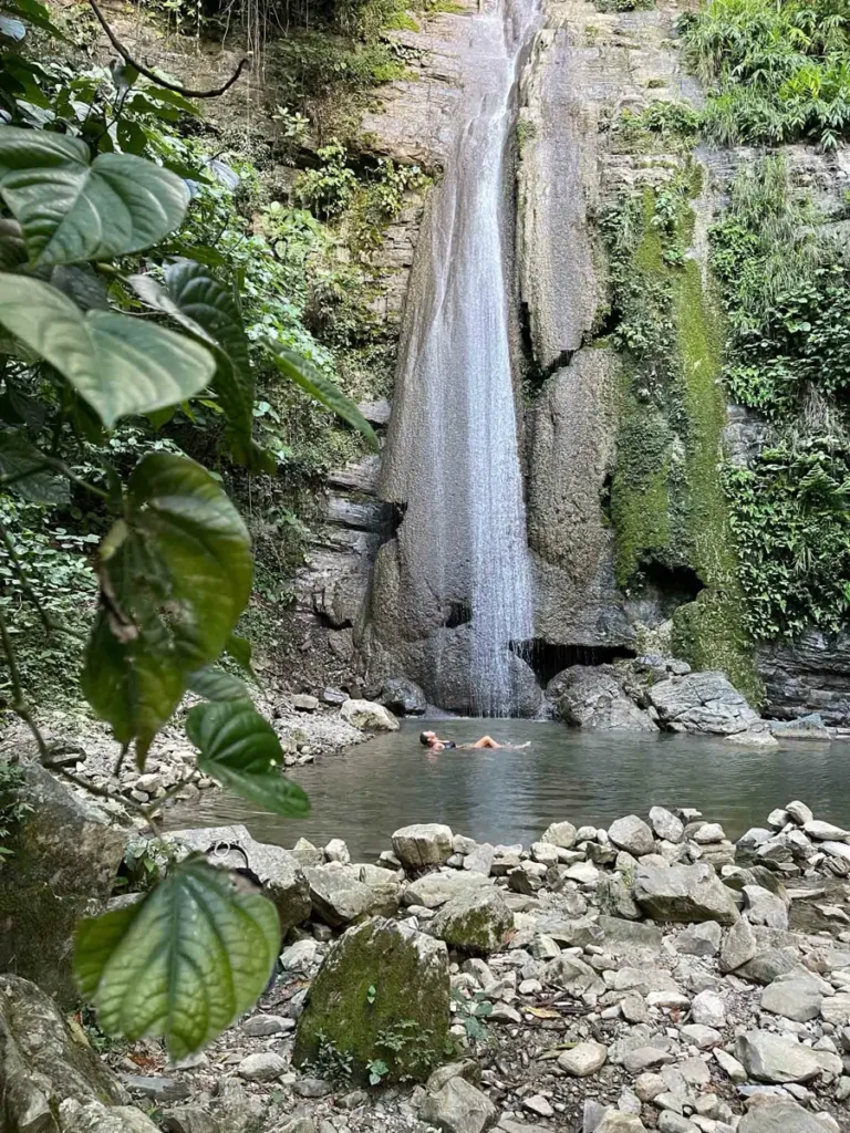hidden no name waterfall in Xilitla with a small body of water and water falling straight from  down and a woman in a black bikini laying in the water surrounded by rocks and a mexican forest, ideal for a perfect mexico itinerary