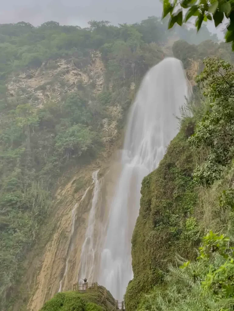 big waterfall Cascada El Chiflon with lots of water streaming down and a woman standing at a viewing platform with a wooden fence stretching her arms to both sides surrounded by green hilly rocky mexican forest