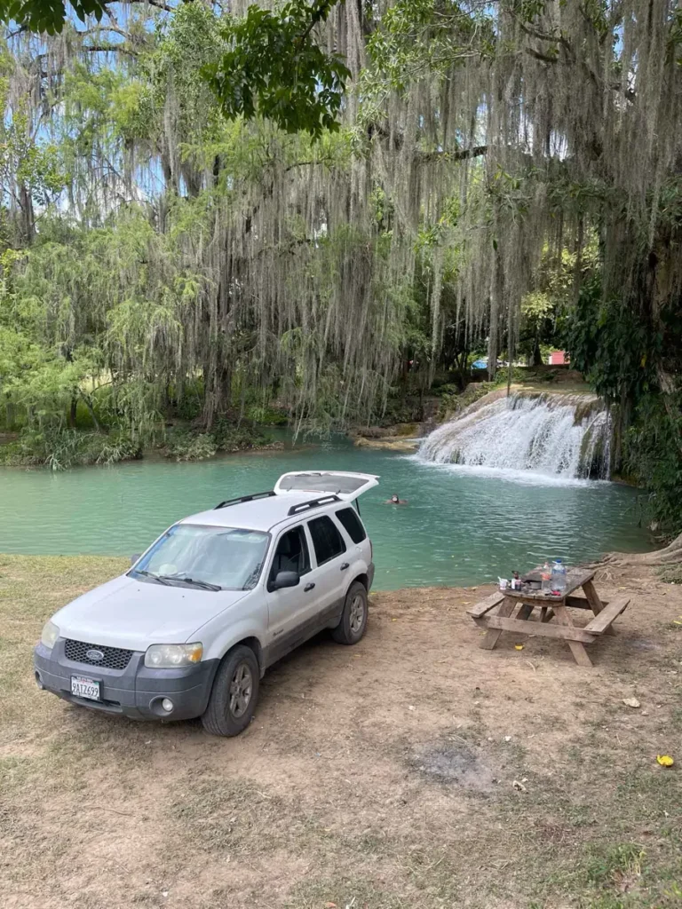 parked white car with an open trunk next to a body of water with a waterfall and small wooden camping bench and table in a forest area
