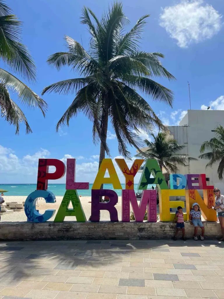 beachside promenade with tiles on the path and a colorful writing playa del carmen with some palm trees and white buildings in the back overlooking the white beach and blue water at playa del carmen in mexico 