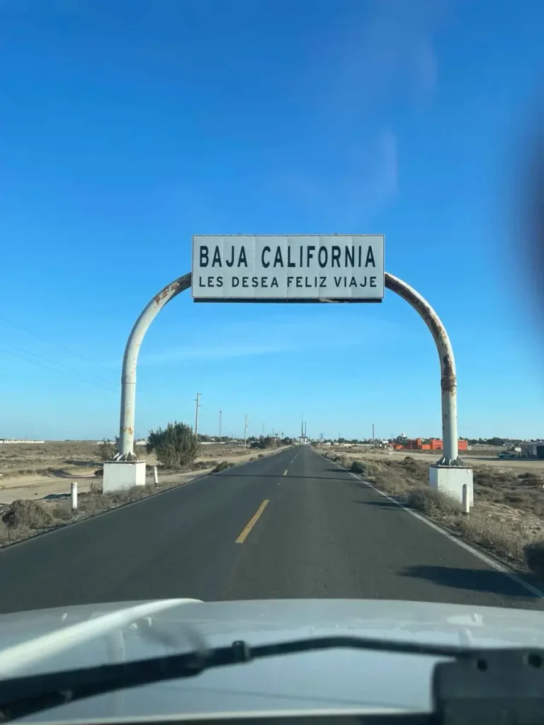 picture from inside the car on a hifhway with a big sign that says baja california in a dry lnadscape