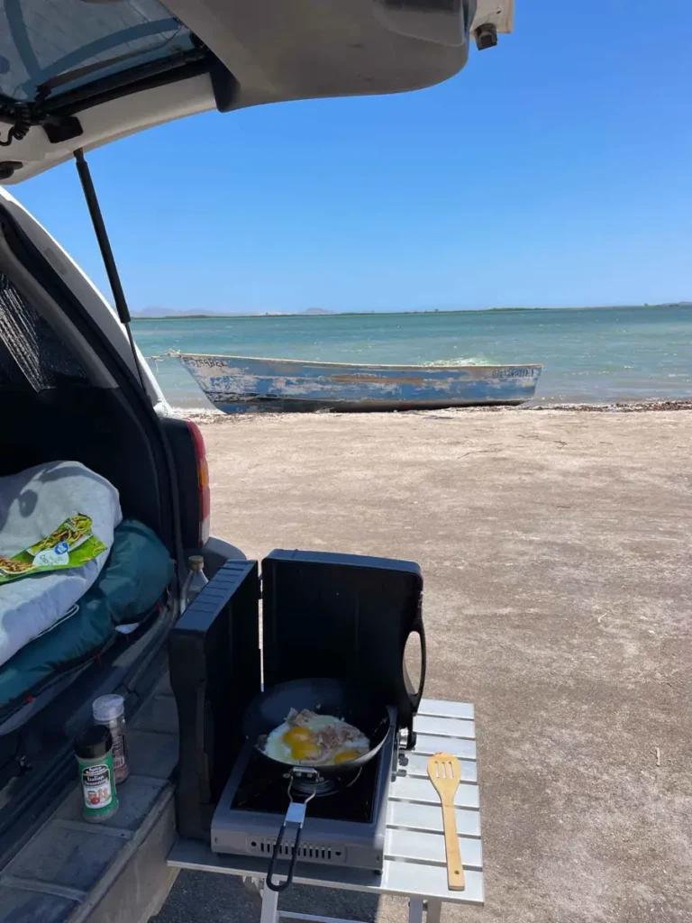 car next to the ocean with a boat in the back and an open trunk with a small camping chair and table next to the car