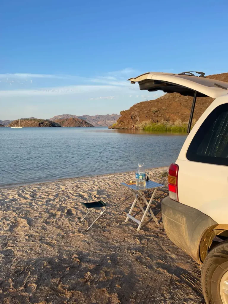 car with an open trunk on a beach with a dry hilly landscape in the back with a camping chair and table next to the white car
