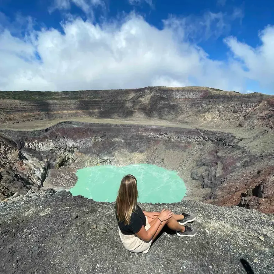 woman sitting on the edge of a volano crates with a view of the steaming bright turquoise crater lake