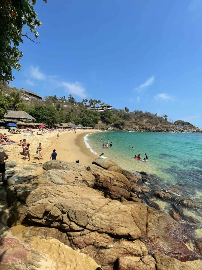Playa Carrizalillo in Puerto Escondido with turquoise water and some people on the beach with beiga rocks surrounded by mexican forest area and a few huts and houses in the back