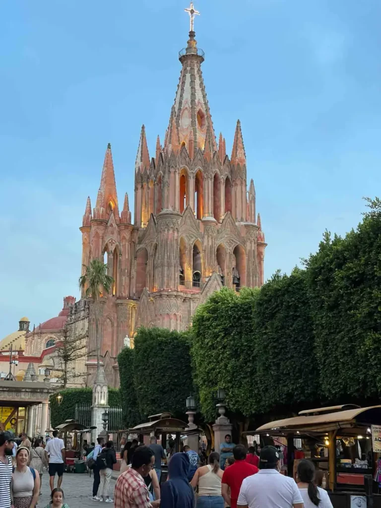 view from the street of the cathedral San Miguel de Allende with many details of arches and spikes and some lights inside the cathedral next to a high hedge and a pedestrian street with small vendors
