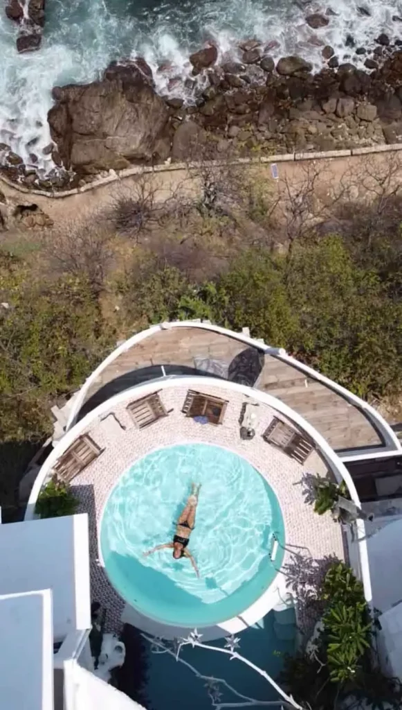 pool with a view at Hotel Pasion de Luna in Mexico woman in a black bikini floating in a small round blue pool surrounded by wooden lay chairs and tables at a white house with a unique shape and multiple teraces surrounded by mexican nature next to an path next to the sea where waves hit some rocks at Hotel Pasion de Luna in Puerto Angel