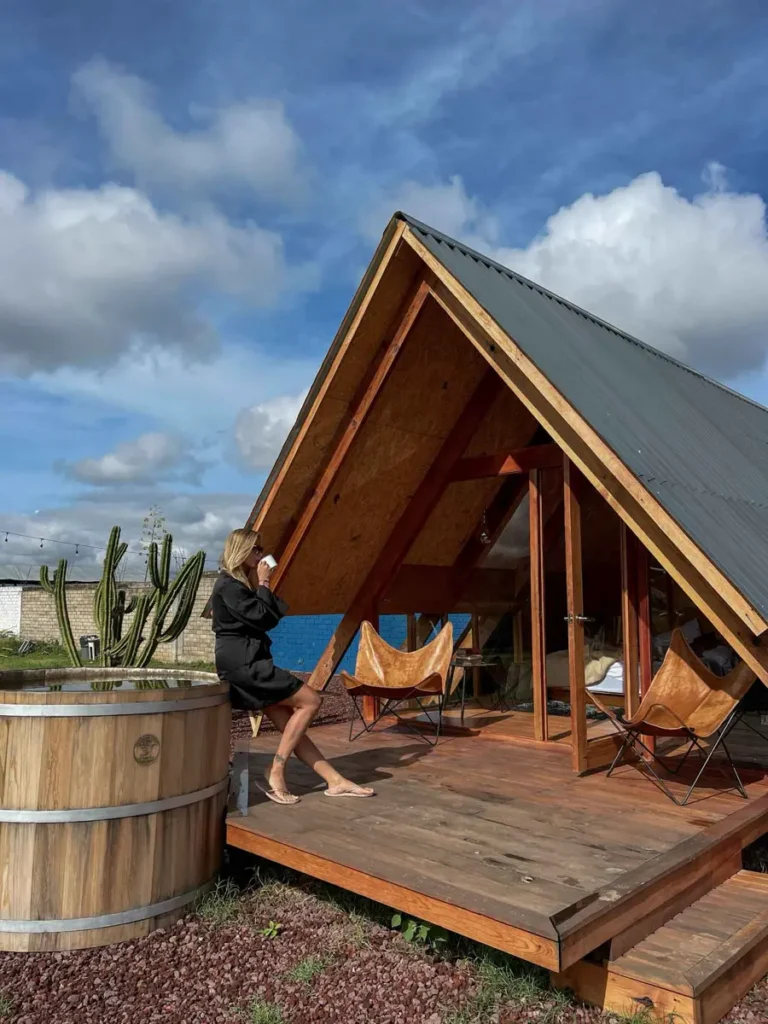 unique places to stay in Mexico: A-frame cabin close to Oaxaca City a fram wooden cabin etlan cabana in mexico with a wooden platform and wooden hot tub with leather chairs and a woman in a black outfit in front of the cabin surrounded by mexican nature and a big cactus