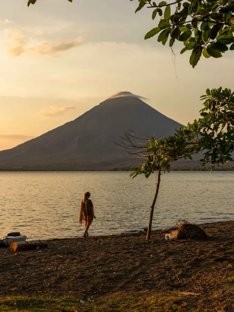 woman walking on the shores of a volcanic lake during sunset with a volcano in the background and some leaves in the foreground