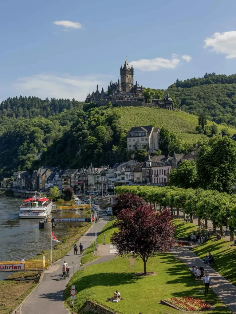 small town along a river with an impressive castle on a hill above the town with boats docking along the shores of the lake and many trees and greenery around