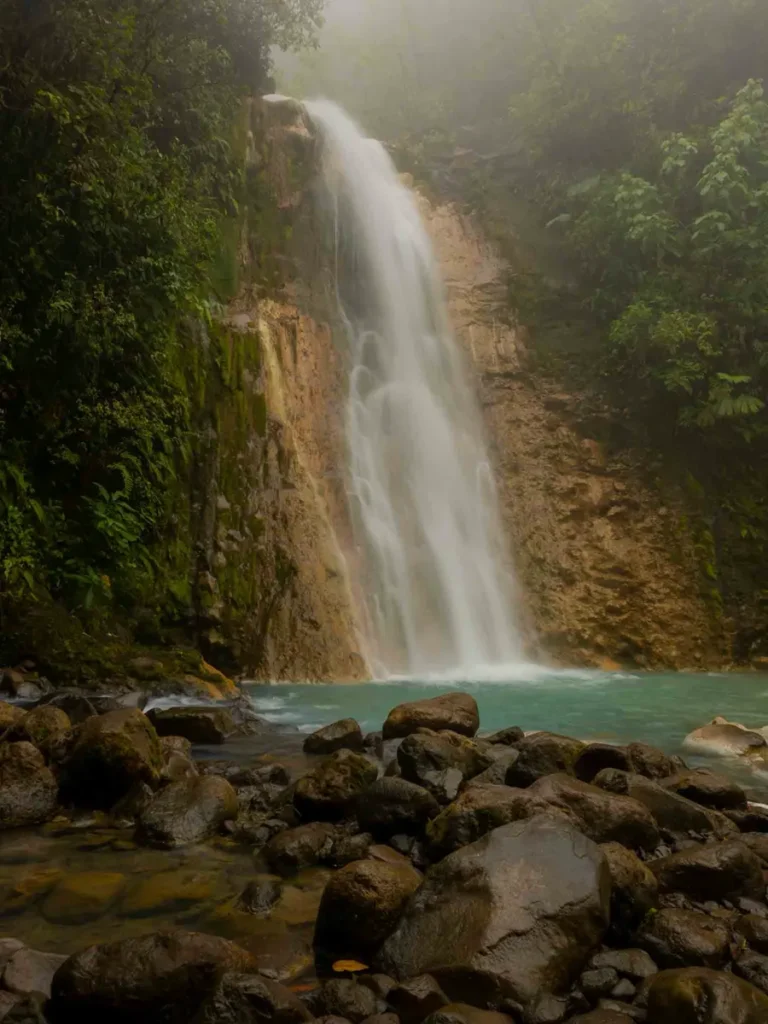 waterfall forming a round blue body of water surrounded by rocks, overgrown green rocks and the jungle with heavy at Blue Falls of Bajos del Toro: Cataratas des Gemelas in costa rica