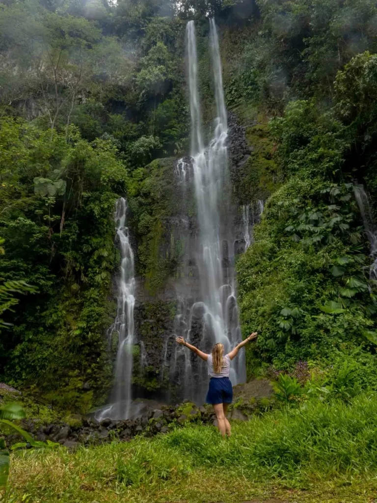 paraiso manatiales waterfall with a blonde woman standing in front of it and stretching her arms to the side surrounded by overgrown rocks and the jungle in bajos del toro in costa rica