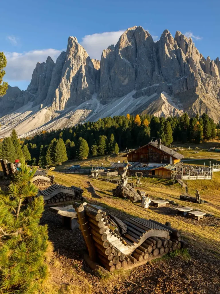Wooden sunbeds on a hill overlooking a picturesque mountain hut with impressive mountain peaks towering above it at Cinema delle Odle in the Dolomites
