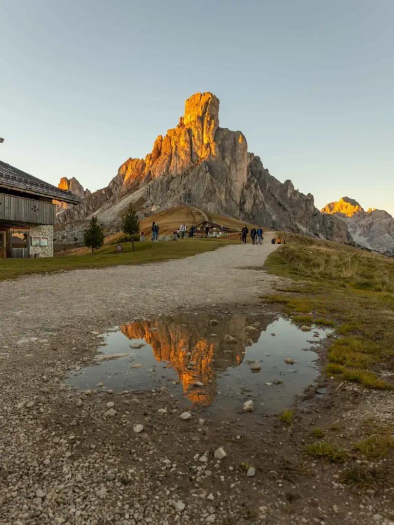 A scanic mountain peak lit up with warm colours during sunset and a reflection of the peak in a puddle on a walking path leading to a mountain hut at Passo Giau in the Dolomites