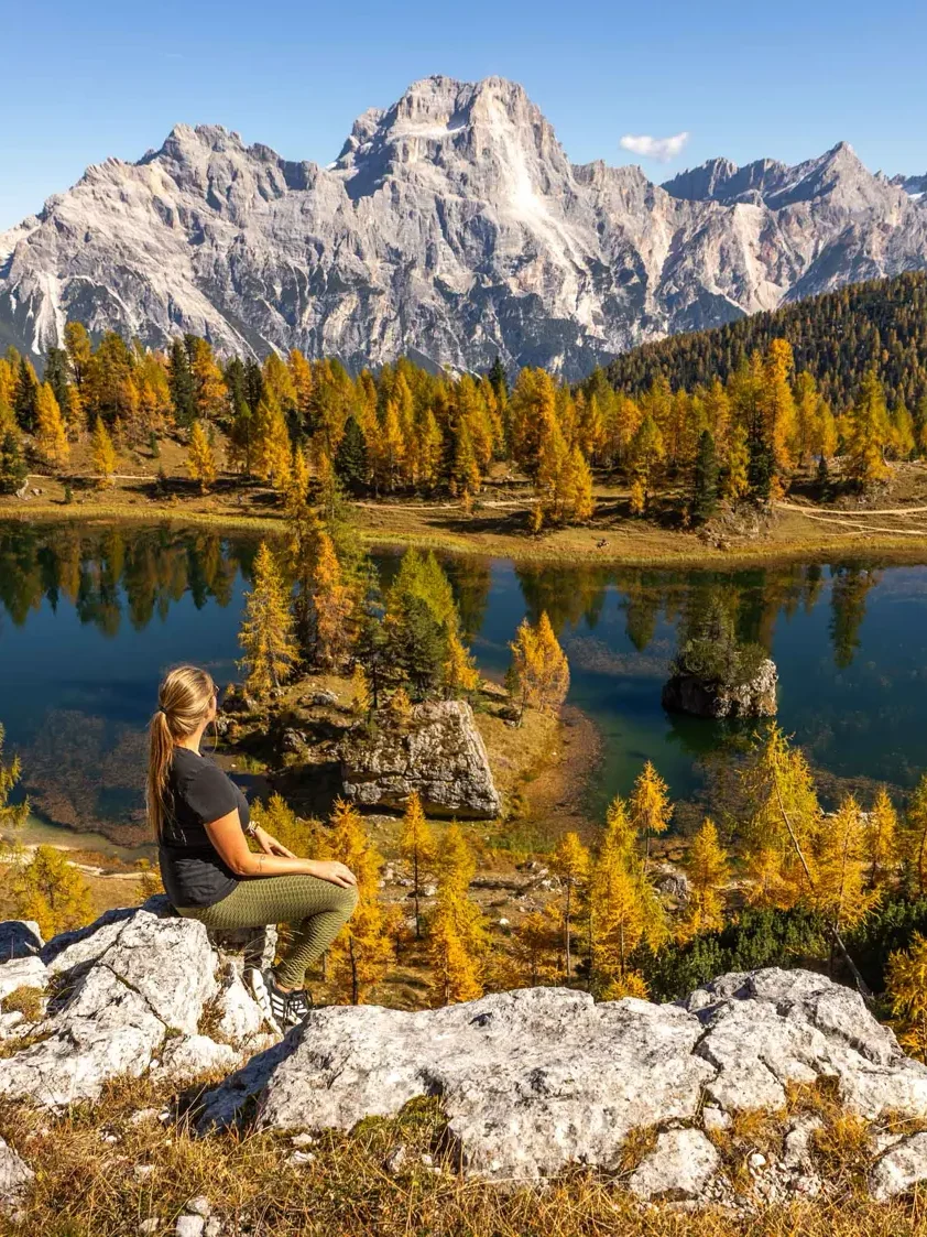 A woman sitting on top of a rock overlooking Lago Federa in the Dolomites with mountain views and golden larches all around
