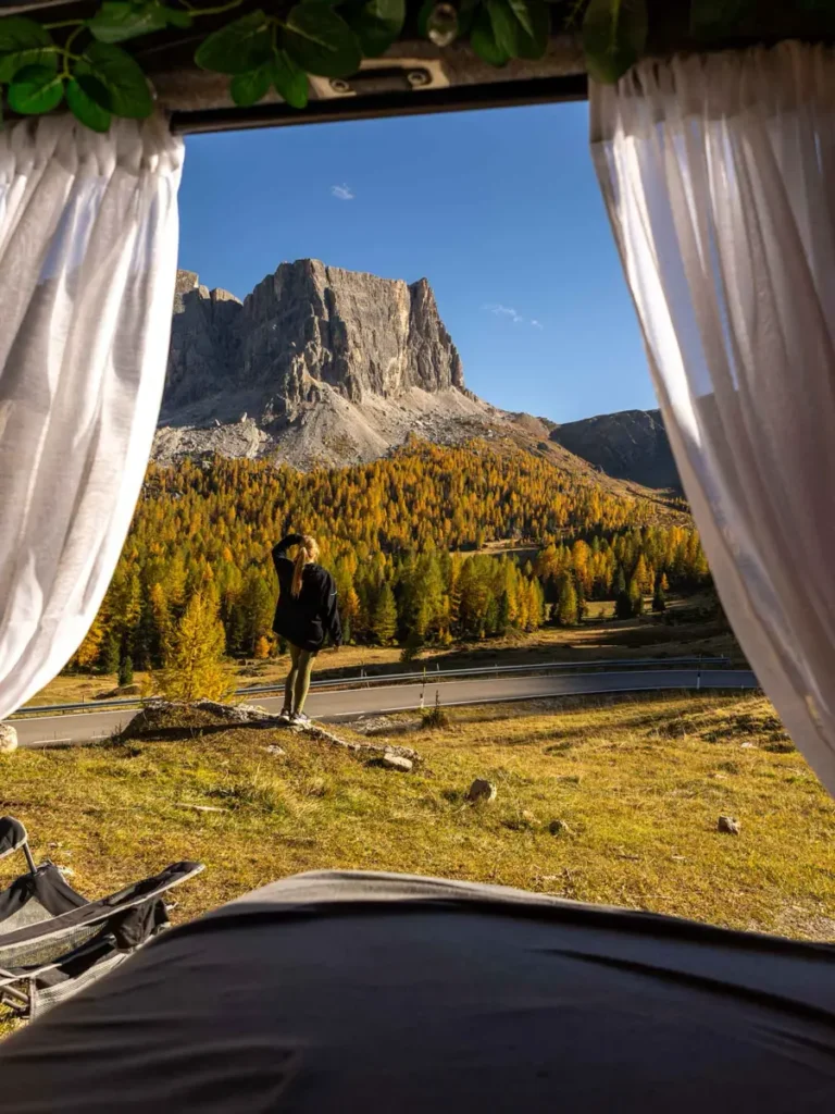 View from the inside of a camper van of a stunning mountain peak towering above golden larches and a woman enjoying the view during autumn in the Dolomites