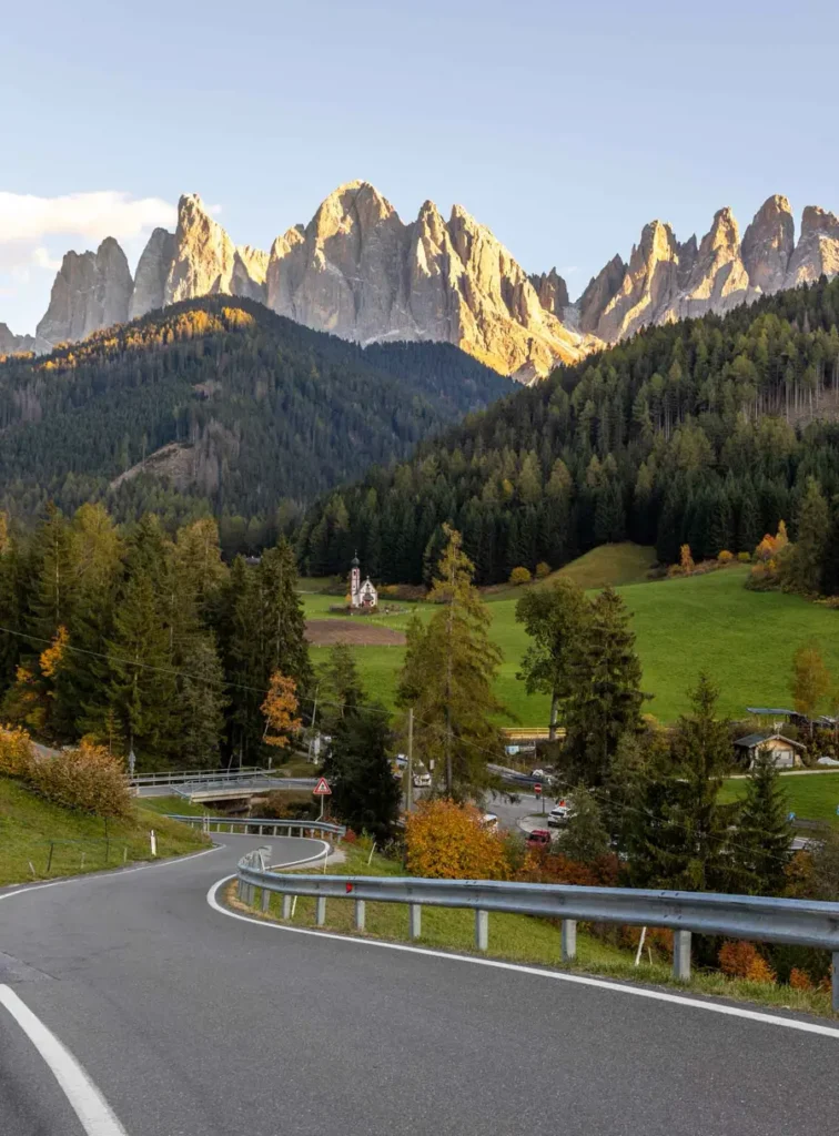 A road leading down a hill to a lonely church on green lawn with towering peaks in the backdrop in Val di Funes, Dolomites