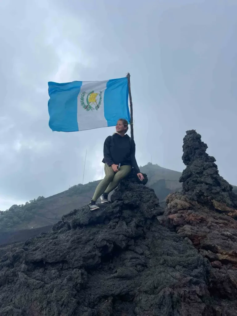 blonde woman in a black outfit sitting on volcanic rocks next to the guatemalan flag at the pacaya volcano hike in antigua guatemala in black volcanic landscape