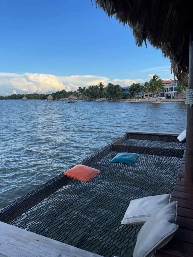 net to lay in over the water with pillows and next to a bungalow at the sea with a view of some hotels at the beach and palm trees and bungalows on the water at Big Dock at Jaguar Reef in hopkins belize