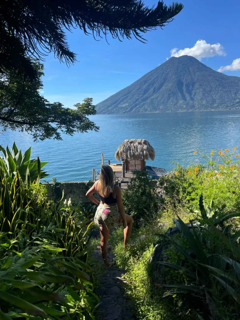 blonde woman standing looking at lake atitlan, a small wooden dock with a hut and a volcano in the back standing in between different plants and bushes at lake atitlan in guatemala