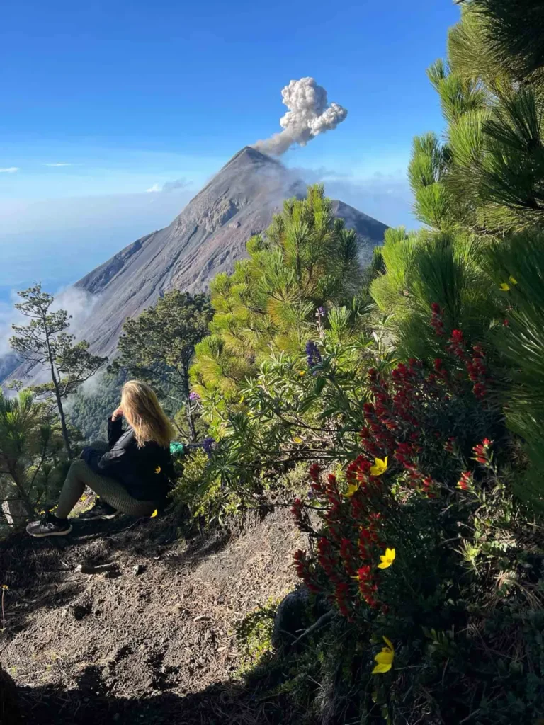 blonde woman in a black outfit sitting during the Acatenango hike with views of erupting volcano Fuego surrouned by black volcanic landscape and some greenery in antigua guatemala