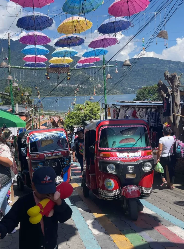 two tuk tuks at the small art town San Juan La Laguna at lake atitlan with cobble stones, small houses, many tourists and colorful umbrellas hanging over the street with some views on the lake and volcanoes at lake atitlan in guatemala