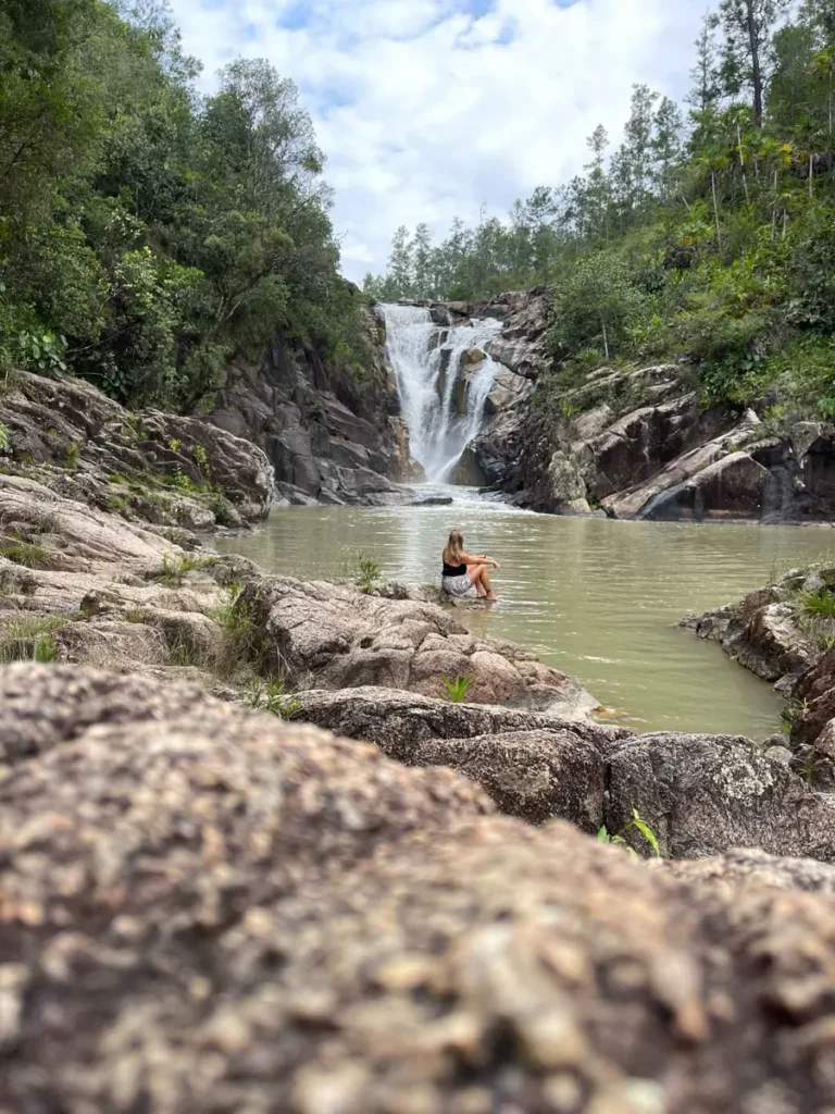 woman sitting next to a body of water with a waterfall in the back surrounded by rocky landscape and a pine forest at Big Rock Falls at mountain pine ridge reserve in belize