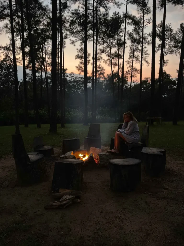blonde woman in a grey cardigan sitting on a tree trunk building a circle with multiple more around a campfire surrounded by pine trees at sunset at mountain pine ridge in belize