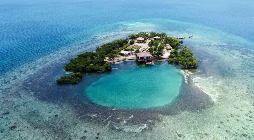 small island with greenery and some housing including overwater bungalow surrounded by blue clear water and small blue hole, one of the best hidden gems in Belize: private island rental in Belize - Little Peter Oasis