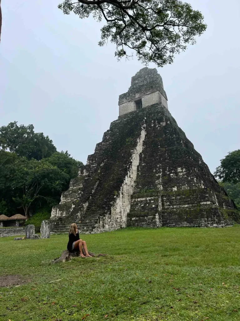 woman in a black outfit sitting in green grass in front of a maya ruin with many stairs forming a pyramid shape at tikal ruins in guatemala