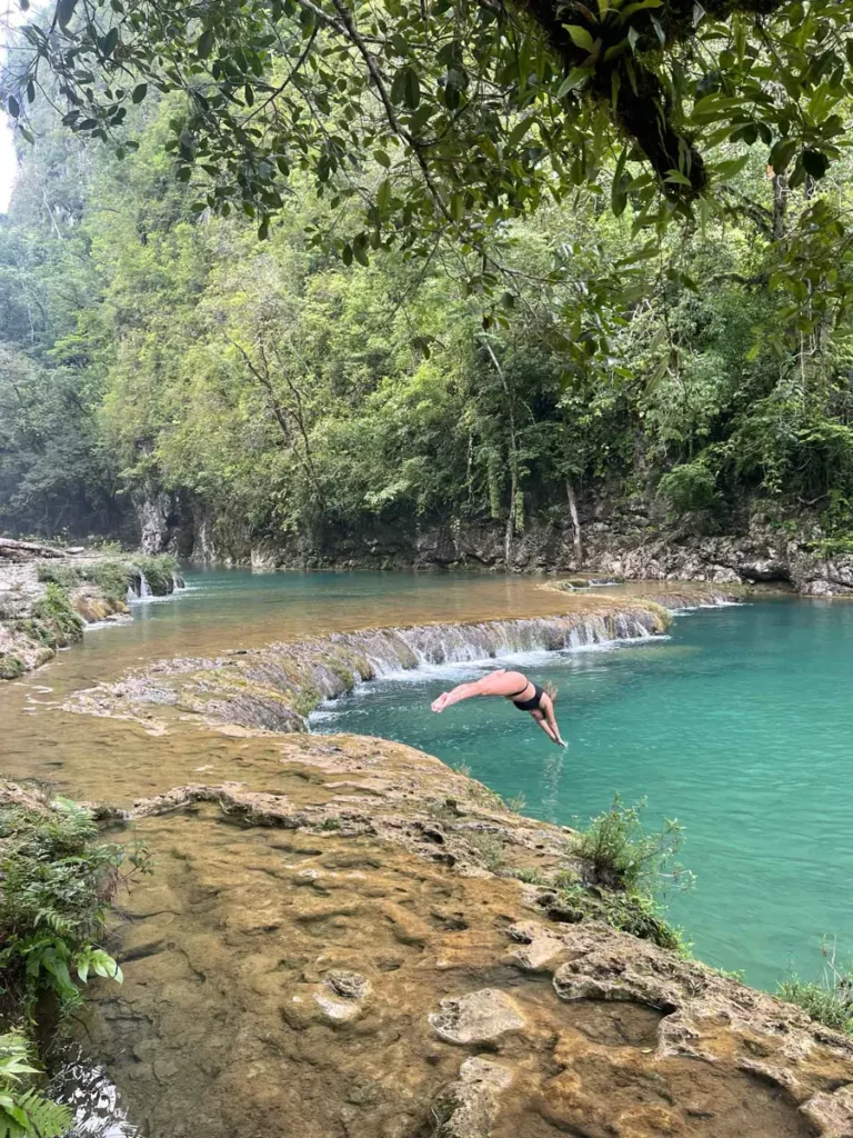 blonde woman in a black bikini jumping in the clear and turquoise water at semu champey a river with multiple small waterfalls and pools with overgrown rocks in the jungle in guatemala