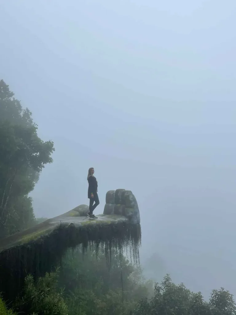 woman in a black outfit standing on a big stone man made hand which is overgrown by some plants in heavy fog in hobbitenango in antigua guatemala