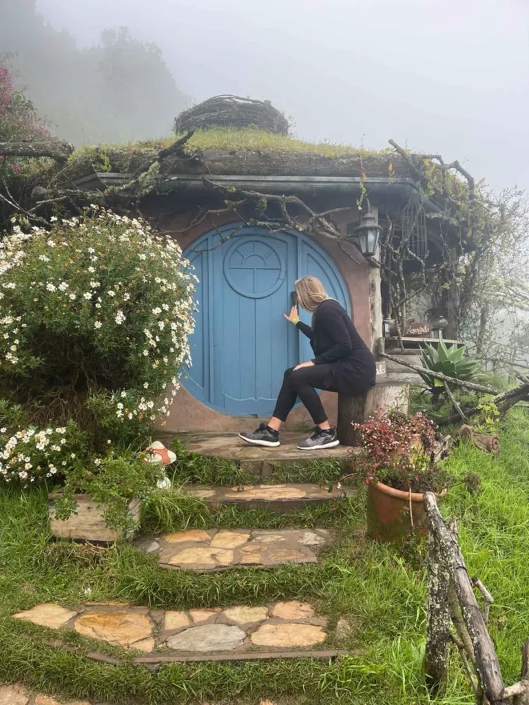 blonde woman in a black outift sitting in front of a small blue hobbithouse with a few steps leading to the house, the roof overgrown and some bushes around in hobbitenango in antigua guatemala