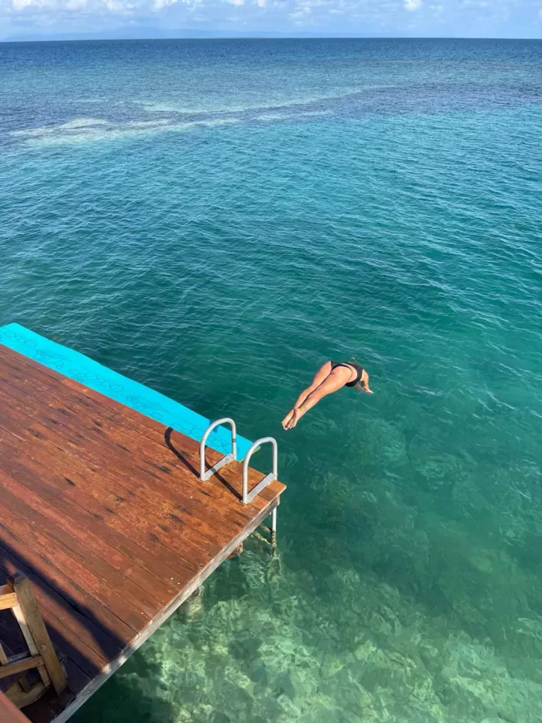 woman in a black bikini jumping into the blue ocean from a wooden gate with a ladder at little peter oasis, a private island rental in belize