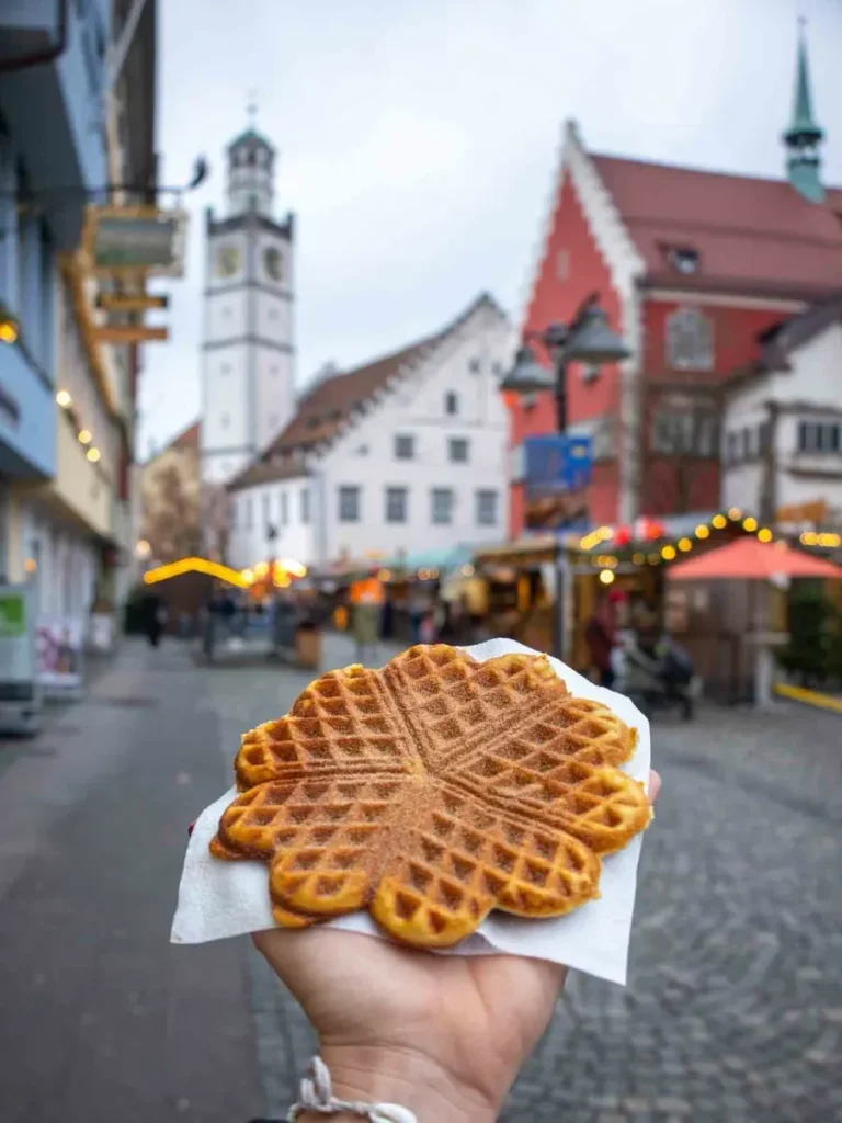 Hand holding a waffle at a Christmas market with stalls and a view of a church in Ravensburg