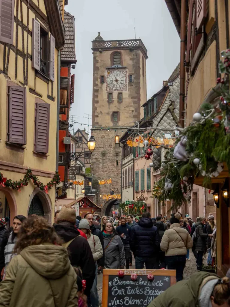 Cobblestone road filled with pedestrians and Christmas decorations leading to an historic clock tower in Ribeauvillé