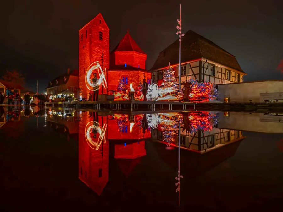 A fire show on a stage on the water with reflections of the artists, church and a traditional Alsacian house in Ottmarsheim