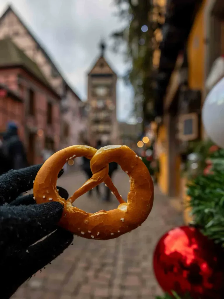 Hand holding a bretzel with colourful houses, christmas decorations and a clock tower in the background in Riquewihr, Alsace