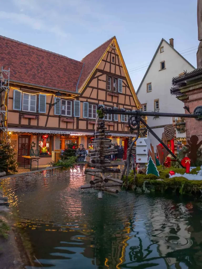 A traditional yellow half-timbered house in Eguisheim with a fountain with reflections in the foreground